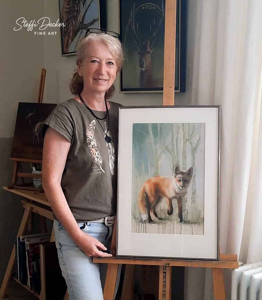 Steffi Decker Steffi Decker steht in einem Atelier neben einer Staffelei und hält ein gerahmtes Gemälde eines Fuchses vor einem Waldhintergrund; im Raum hängen weitere Tiergemälde an der Wand und Malutensilien stehen auf einer Holzstaffelei.